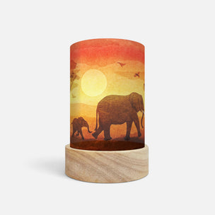 A cylindrical table lamp with a light wood base and a printed wraparound cylinder body featuring a sunset scene with elephants, birds, and a large sun, shown on a plain light gray background.