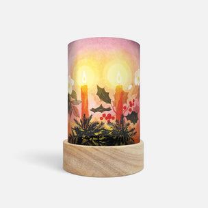 A cylindrical table lamp with a light wood base and a printed wraparound shade depicting two glowing candles, holly leaves, berries, and a soft pink-to-orange sunset background.