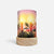 A cylindrical table lamp with a light wood base and a printed wraparound shade depicting two glowing candles, holly leaves, berries, and a soft pink-to-orange sunset background.
