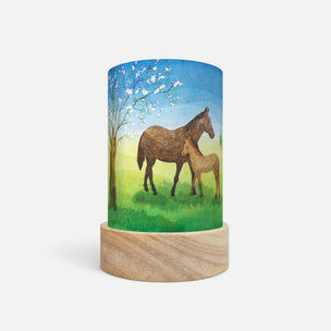 A cylindrical table lamp with a light wood base and a printed wraparound cylinder featuring a painted scene of a brown horse and foal under a blue sky with white blossoms, shown on a light gray background.