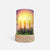 A cylindrical candle holder with a printed illustration of three lit candles, evergreen branches, and a sunset sky, set on a round light wood base against a light gray background.