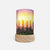 A cylindrical table lamp with a light wood base and a printed wraparound cylinder featuring four lit candles in front of a sunset sky with palm tree silhouettes, shown on a plain light gray background.