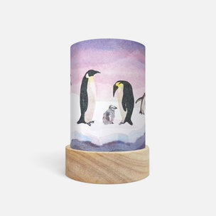 A cylindrical lamp or candle-style decor piece with a watercolor illustration of penguins on a snowy landscape, mounted on a round light wood base against a clean white background.