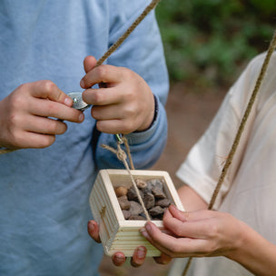 Two children outdoors, one holding a small wooden box with stones and a string, the other assisting.