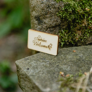 Small wooden sign on a stone surface with moss, featuring the text 'Faires Welcomes'.