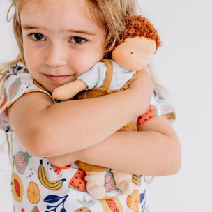 A young child with blonde hair hugging a soft plush doll with brown hair, wearing a white fruit-patterned shirt against a plain white background.