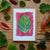 Card with leaf design on pink background surrounded by real leaves on wooden surface