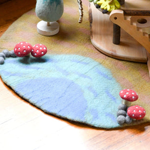 A handcrafted felt mushroom-themed play rug on a wooden floor, featuring a blue and green leaf-shaped base with red and white felt mushrooms at the corners and small gray felt mushrooms along the edges.