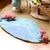 A handcrafted felt mushroom-themed play rug on a wooden floor, featuring a blue and green leaf-shaped base with red and white felt mushrooms at the corners and small gray felt mushrooms along the edges.