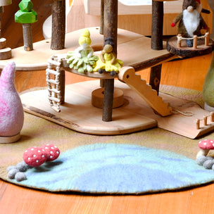 A wooden fairy house playset with a multi-level treehouse structure made of natural wood poles and platforms, featuring small felted wool gnomes, a pink felted wool bird, and a blue felted wool rug on a wooden floor.