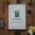 Card with dandelion illustration and text on a wooden surface with flowers