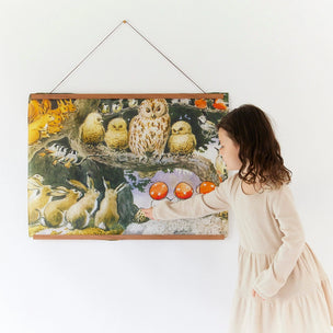 A child in a cream dress stands beside a large framed silk wall hanging depicting owls, rabbits, and photographed against a plain white wall.