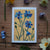 Floral painting with blue flowers on a yellow background, surrounded by real flowers on a wooden surface.