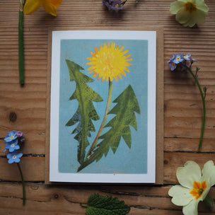 Card with a dandelion illustration surrounded by real flowers on a wooden surface