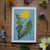 Card with a dandelion illustration surrounded by real flowers on a wooden surface