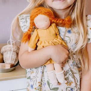 A young girl in a floral dress holds a handmade cloth doll with bright orange yarn braids and a mustard yellow dress.