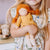 A young girl in a floral dress holds a handmade cloth doll with bright orange yarn braids and a mustard yellow dress.