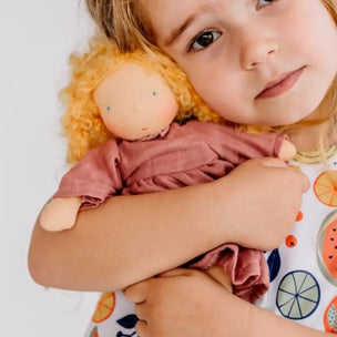 A young child with blonde hair holds a soft plush doll with curly yellow hair and a dusty rose dress against a clean white background.