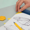 Hand placing a yellow geometric shape on a design sketch with a pen on a light green surface.