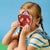 A young child with braided hair holds a bear-shaped mirror in front of their face against a bright blue background.