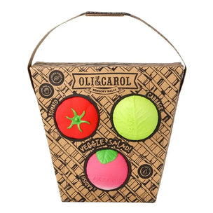 Oli & Carol Sanitary Balls in a brown patterned storage box with a carrying handle, featuring three colorful balls: a red tomato-shaped ball, a lime-green cabbage-shaped ball, and a pink avocado-shaped ball.