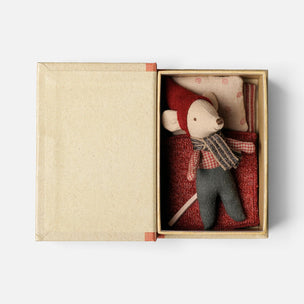 A small Maileg mouse toy dressed in a red hat and scarf with gray pants, nestled inside an open kraft paper gift box with a red patterned blanket and pillow.
