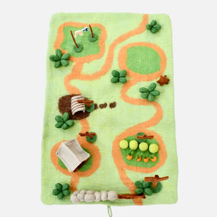 A felted wool activity rug shaped like a golf course map, featuring green grassy areas, orange winding paths, clover shapes, a small flag, a basket, a golf ball, and a putt-putty, on a light gray background.