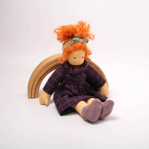 Toy doll with red hair and a purple dress sitting on a wooden rainbow against a white background