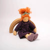 Toy doll with red hair and a purple dress sitting on a wooden rainbow against a white background
