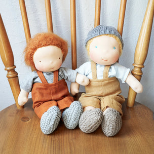 Two handmade cloth dolls sitting side by side on a wooden chair. The doll on the left has curly orange hair, wearing brown overalls and gray knit boots.