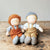 Two handmade fabric dolls sitting side by side on a rustic wooden table against a neutral gray background. The doll on the left has curly orange hair, wearing brown overalls, a gray shirt, and gray knit booties.