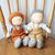 Two handmade cloth dolls sitting side by side on a wooden chair. The doll on the left has curly orange hair, wearing brown overalls and gray knit boots.