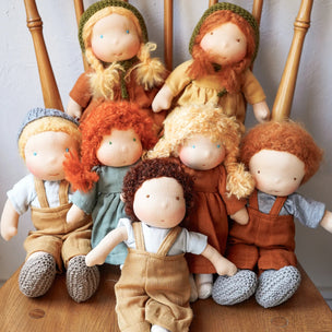 A group of handmade cloth dolls with soft yarn hair and embroidered faces, posed together on a wooden chair against a plain light background.
