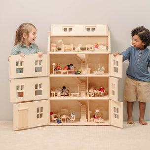 A large wooden dollhouse with open interior rooms and small furniture pieces, shown with two children standing on either side in a clean indoor setting.