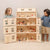 A large wooden dollhouse with open interior rooms and small furniture pieces, shown with two children standing on either side in a clean indoor setting.