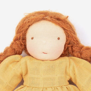 A close-up of a handmade fabric doll with long, curly reddish-brown hair and a textured mustard yellow dress against a plain white background.