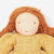 A close-up of a handmade fabric doll with long, curly reddish-brown hair and a textured mustard yellow dress against a plain white background.