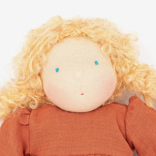 Close-up of a handmade fabric doll with curly blonde yarn hair, a beige stitched face, small blue bead eyes, and an rust-orange dress against a plain white background.