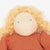 Close-up of a handmade fabric doll with curly blonde yarn hair, a beige stitched face, small blue bead eyes, and an rust-orange dress against a plain white background.