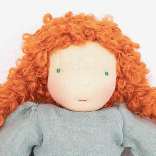 A close-up of a handmade fabric doll with bright orange curly hair, a cream-colored face, small green bead eyes, and a dusty blue-gray dress against a plain white background.