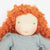 A close-up of a handmade fabric doll with bright orange curly hair, a cream-colored face, small green bead eyes, and a dusty blue-gray dress against a plain white background.