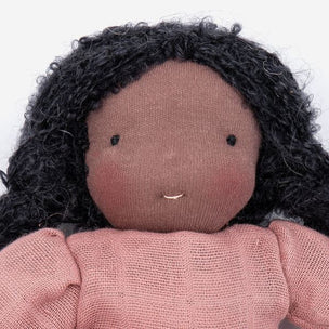 A close-up of a handmade fabric doll with a round dusty pink face, small black embroidered eyes and mouth, black curly yarn hair, and a dusty pink knitted top against a plain white background.