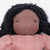 A close-up of a handmade fabric doll with a round dusty pink face, small black embroidered eyes and mouth, black curly yarn hair, and a dusty pink knitted top against a plain white background.