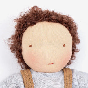 A close-up of a handmade knitted doll with curly brown hair, a beige face, embroidered eyes, and a light gray top with brown suspenders.