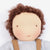 A close-up of a handmade knitted doll with curly brown hair, a beige face, embroidered eyes, and a light gray top with brown suspenders.