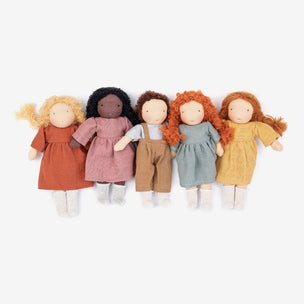 Five handmade fabric dolls with soft cloth bodies and long hair in blonde, black, brown, red, and auburn, dressed in rust, dusty pink, blue-gray, and mustard yellow outfits, arranged side by side on a light gray background.