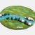 Felted landscape with green grass, blue water, and gray rocks on a white background