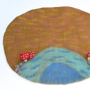 A handcrafted felted wool art piece shaped like a landscape, featuring a golden-brown sky with pinkish stripes, a blue and teal mountain range, and small red-and-white polka dot mushrooms with wooden stems.