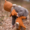 Child in a forest setting wearing a knitted hat and holding a plush toy, surrounded by autumn leaves.