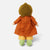 A doll viewed from the back, wearing a mustard yellow knit hat, an orange-brown dress, yellow pants, and light green crocheted shoes, set against a plain light gray background.